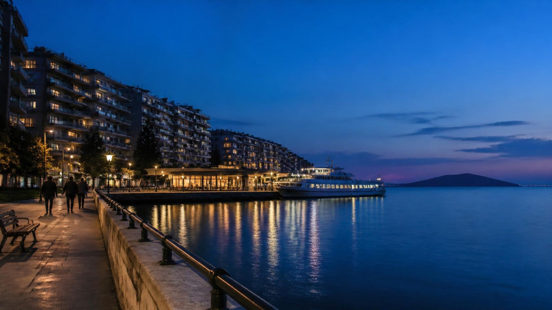 A long stone pier and calm sea at dusk, a passenger boat at a small terminal, and rows of lit apartment blocks along the shore with a low island on the horizon