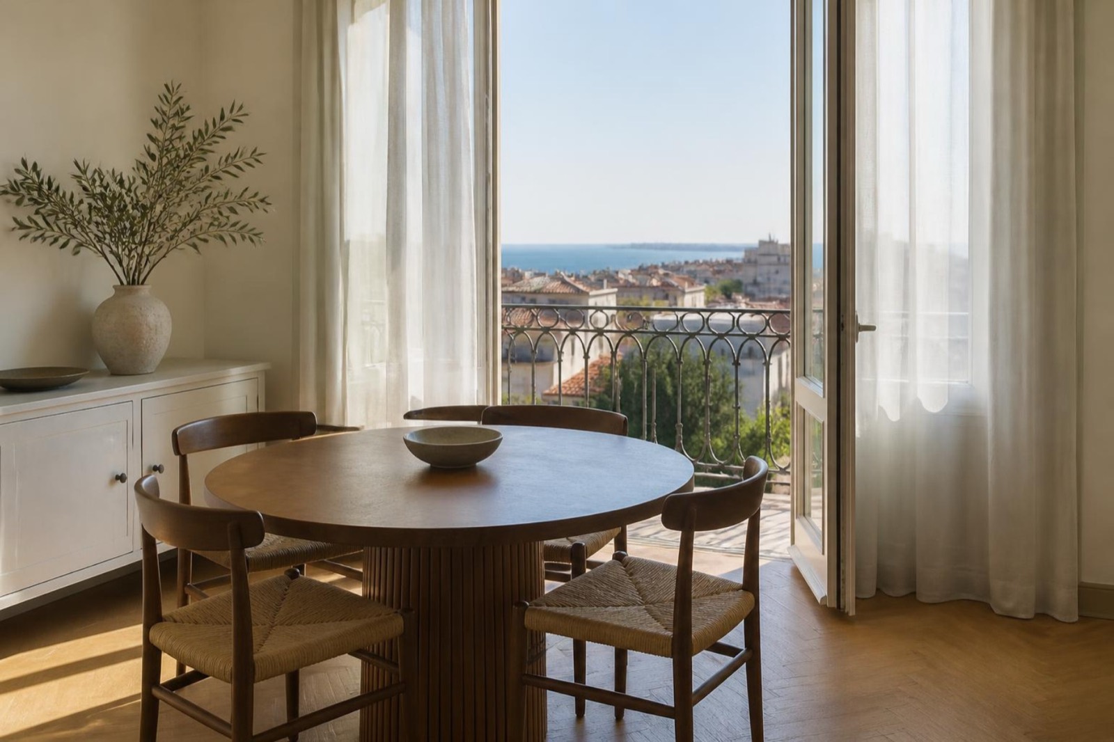 A light dining area with a wooden table beside balcony doors and a strip of blue sea beyond the rooftops