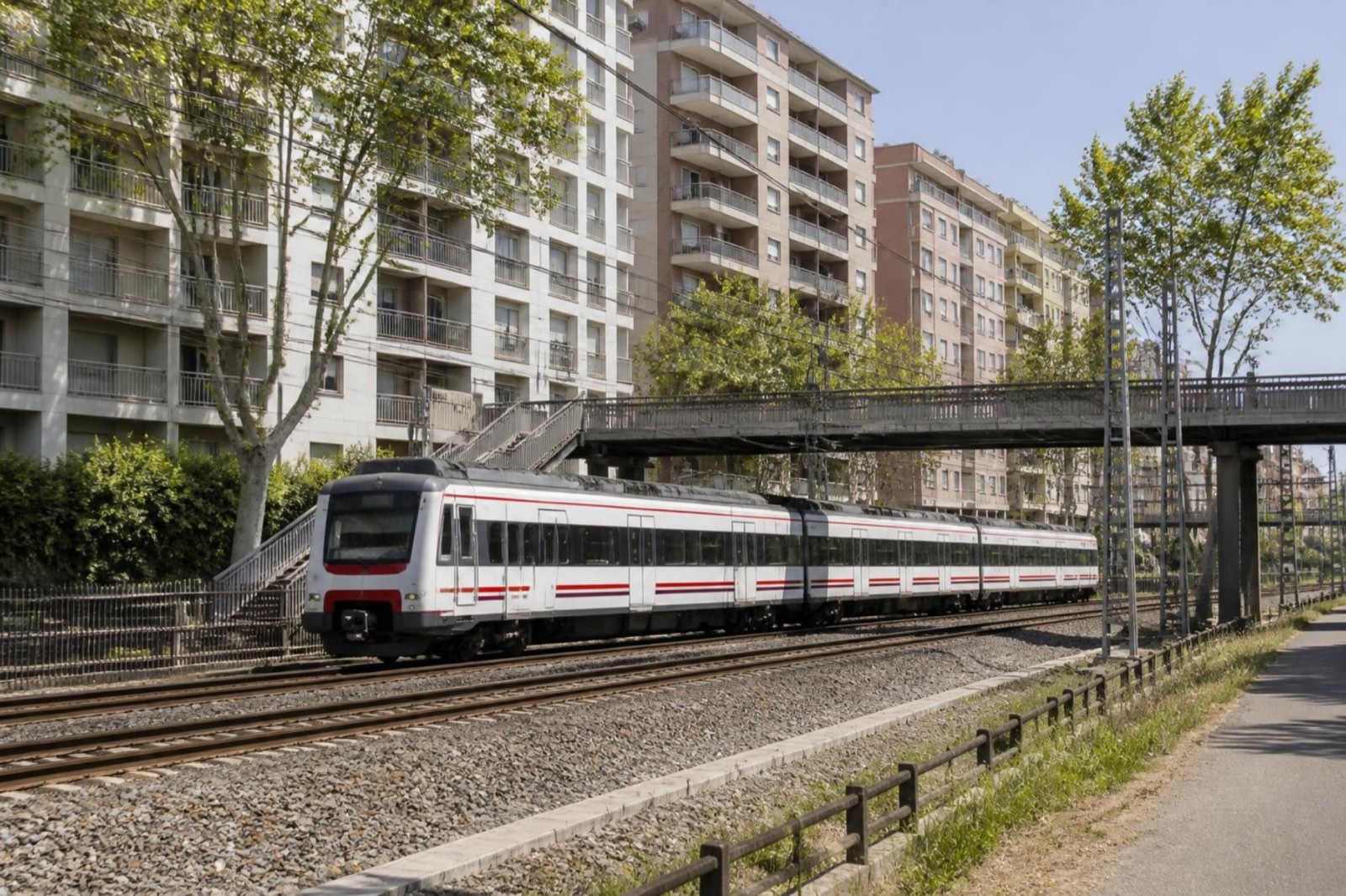 Mid-rise apartment blocks with balconies rising behind a double railway line with overhead wires and a white commuter train